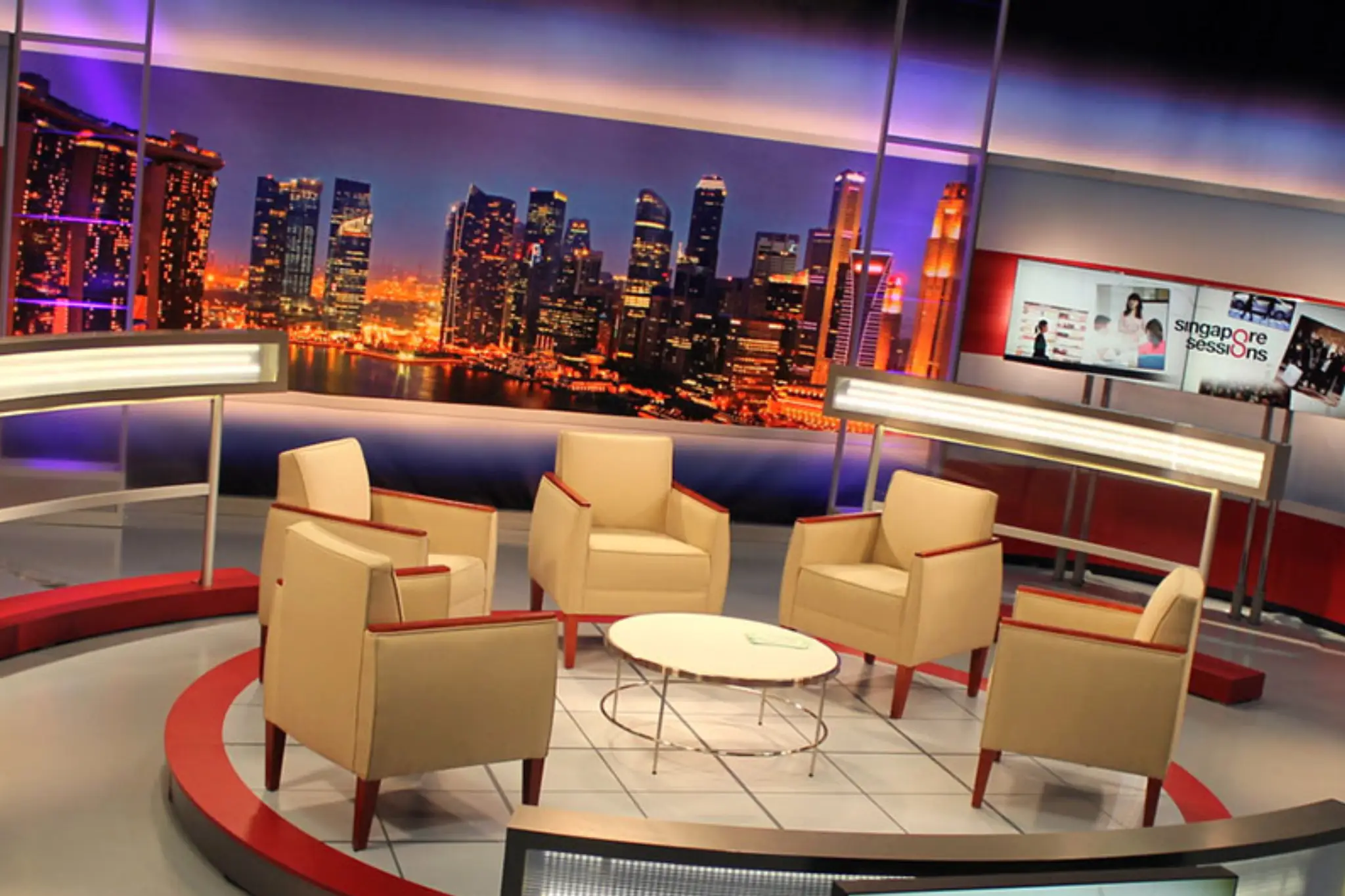 Modern TV studio set with beige chairs and a round table, featuring a vibrant city skyline backdrop and _Singapore Sessions_ display Modern TV studio set with beige chairs and a round table, featuring a vibrant city skyline backdrop and _Singapore Sessions_ display