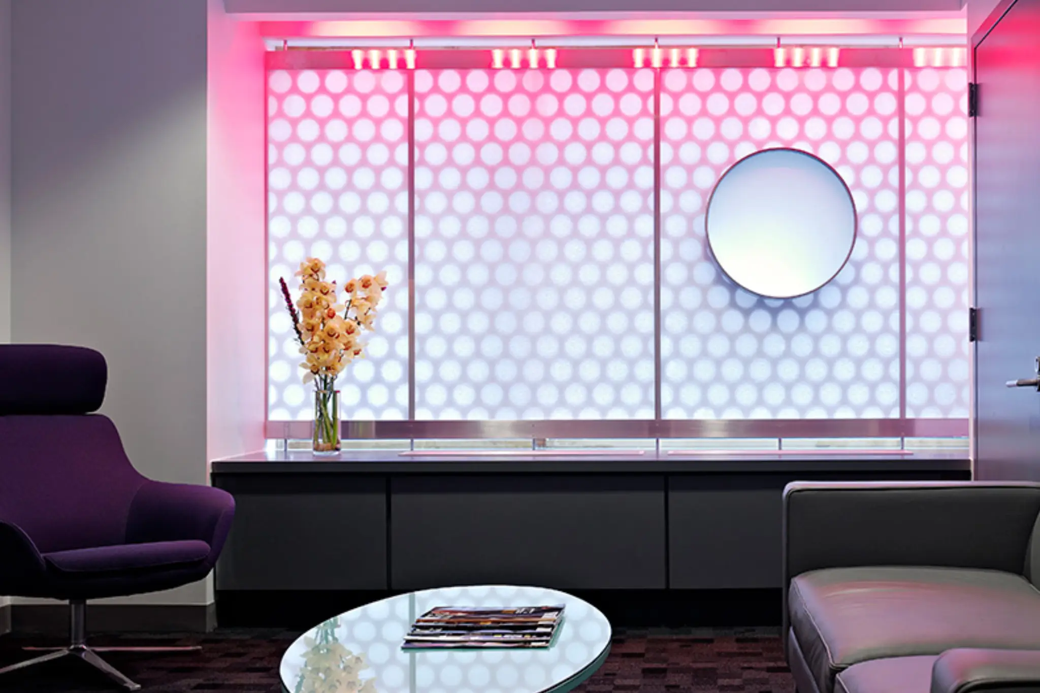 Modern lounge with purple chair, sleek sofa, circular mirror, and illuminated wall displaying patterned lighting. A vase of flowers on a glass table Modern lounge with purple chair, sleek sofa, circular mirror, and illuminated wall displaying patterned lighting. A vase of flowers on a glass table